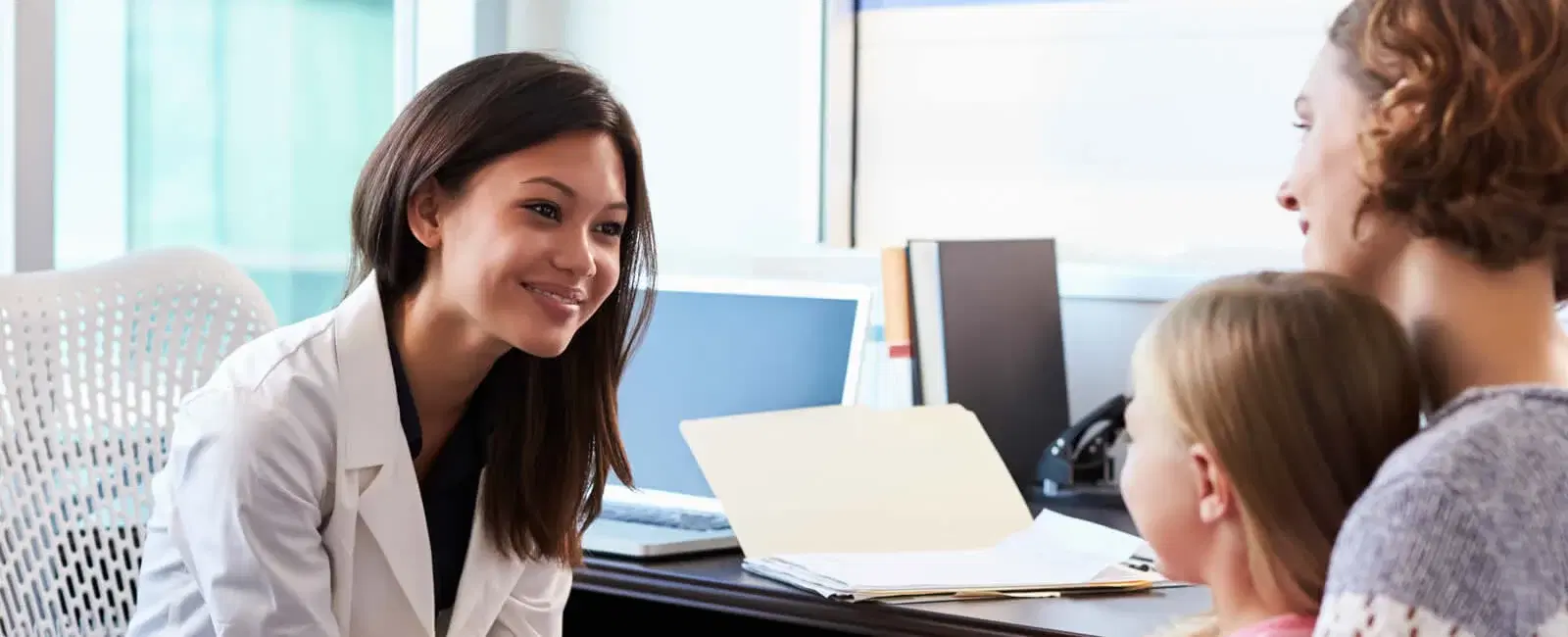 Smiling female doctor talking to a mother and young daughter across a desk in a medical office