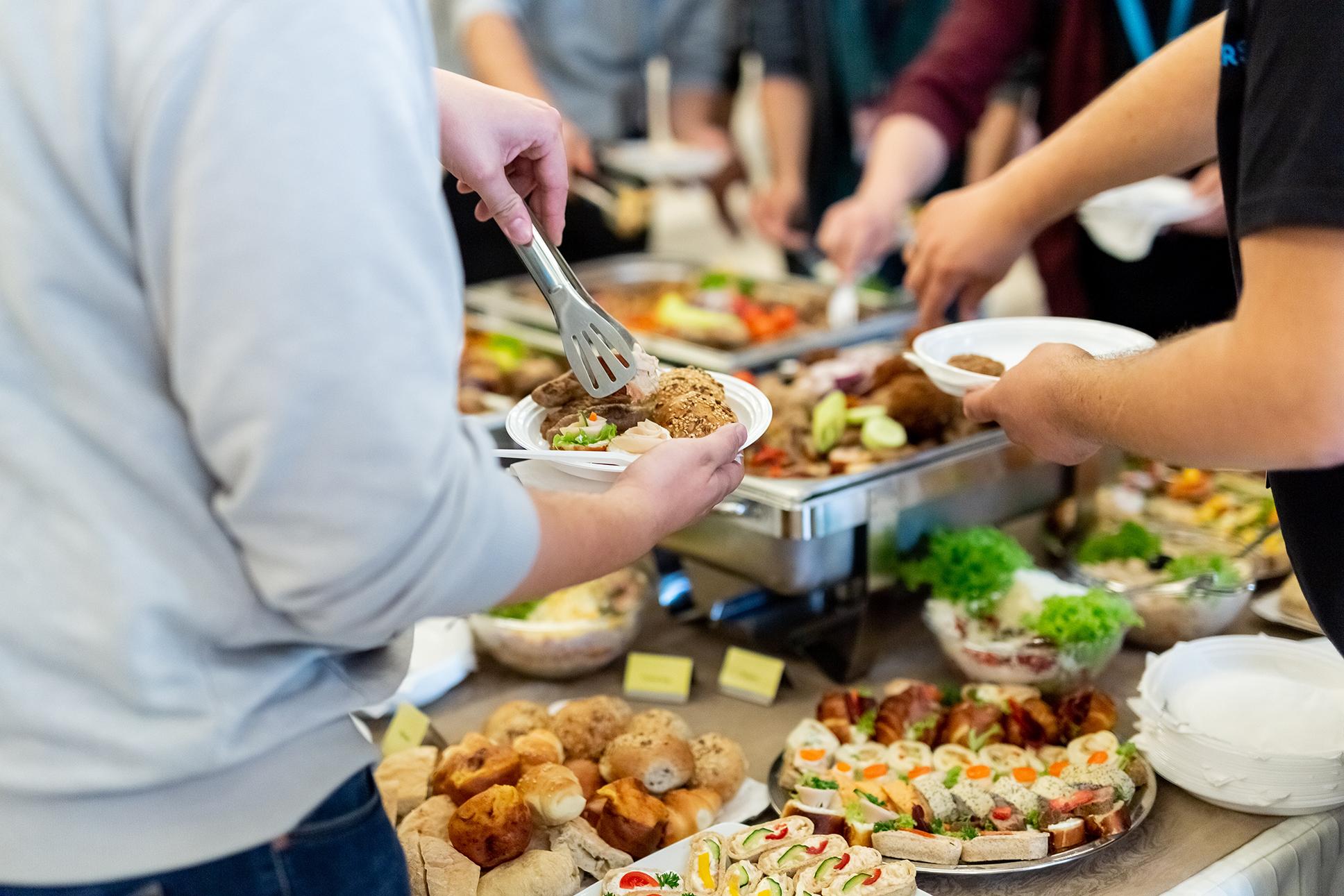 Hands serving rolls and assorted appetizers onto paper plates at a crowded buffet table.