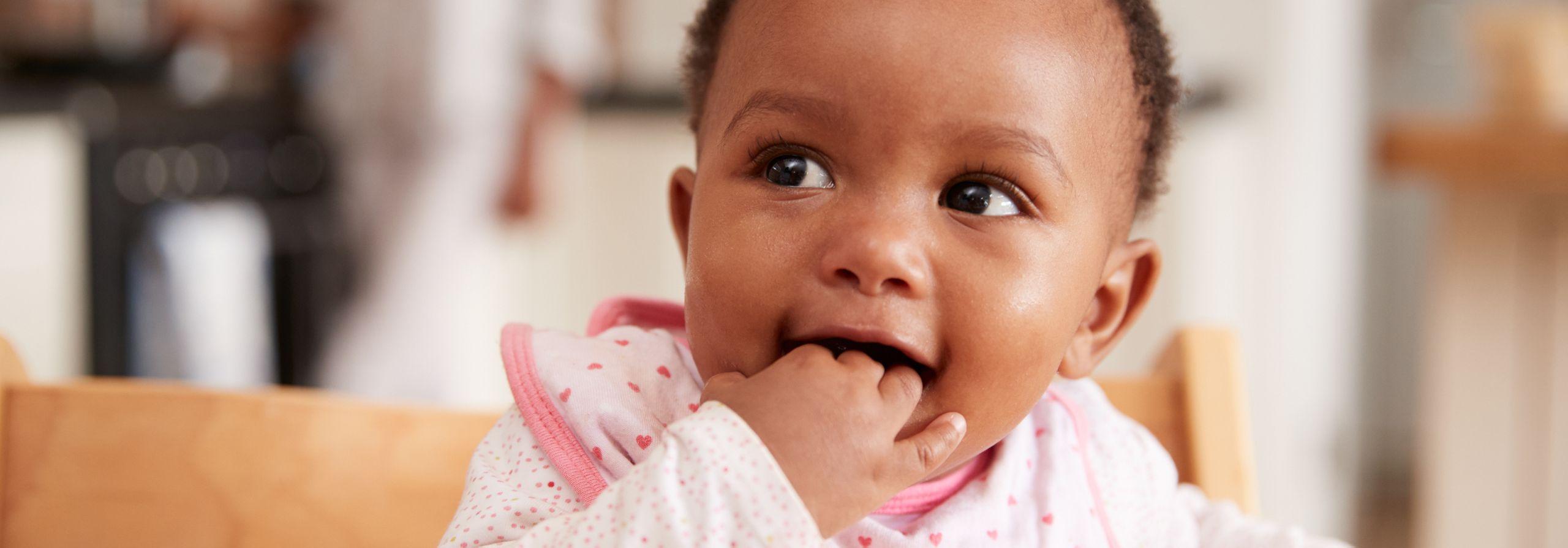 Close-up of a baby in a high chair wearing a pink bib, fingers in mouth and looking off to the side with wide eyes.